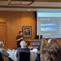 A man holds a microphone in front of a presentation with a video example